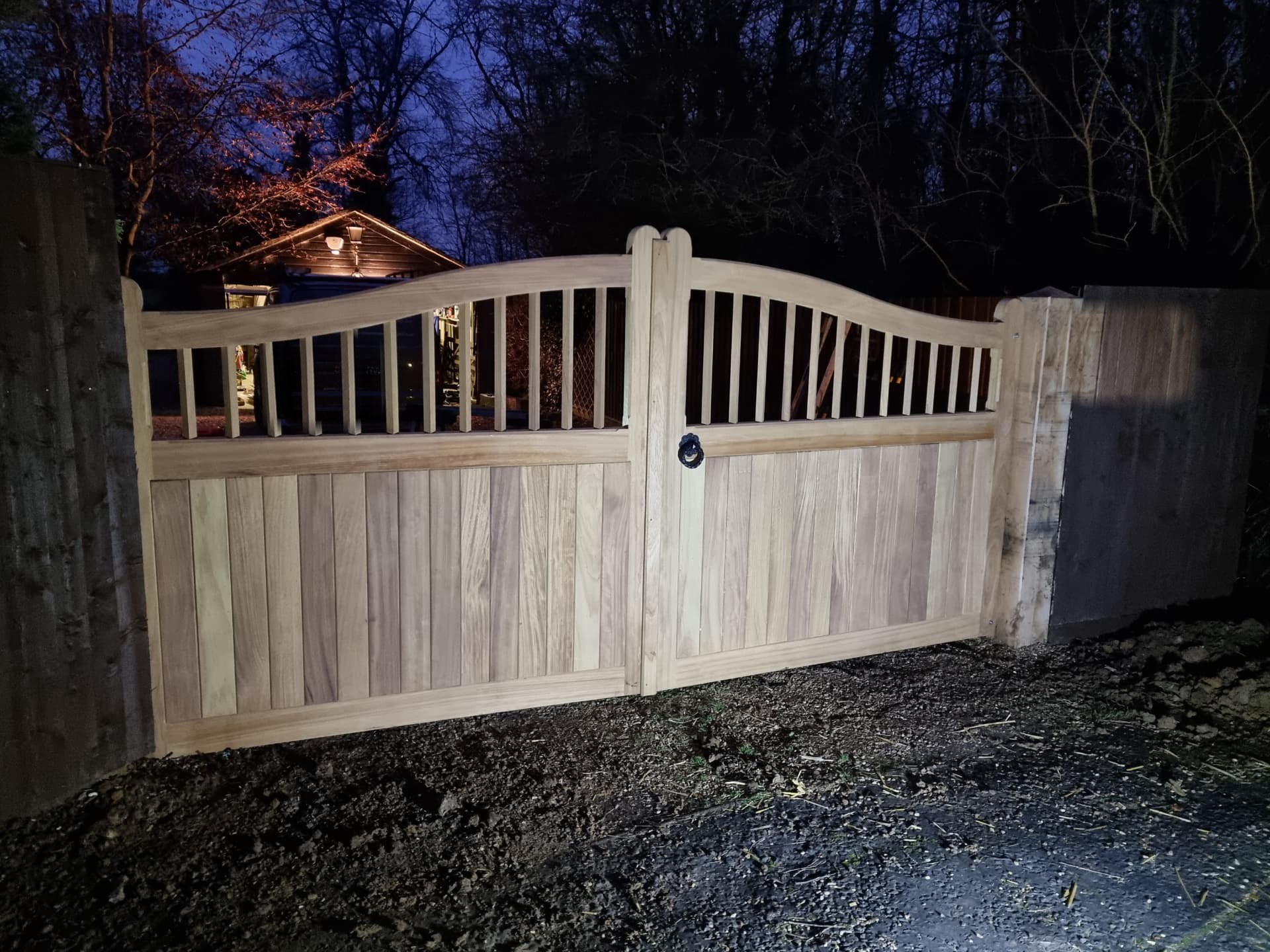 Arched oak driveway gates photographed at dusk, with a curved top and black iron hardware